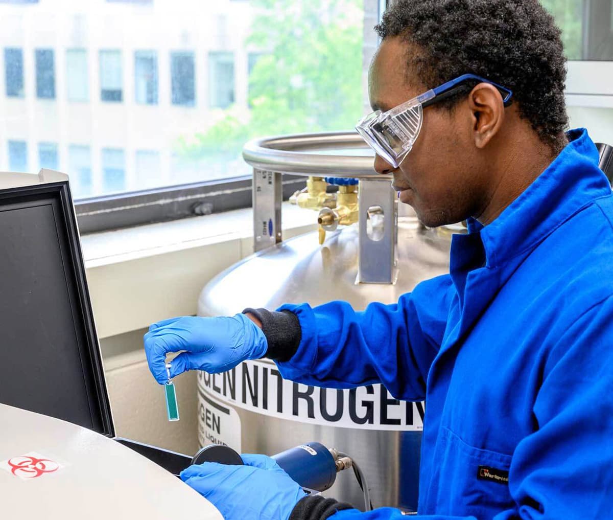 A researcher wearing a blue lab coat, safety glasses, and blue gloves carefully places a small, green sample into a lab instrument